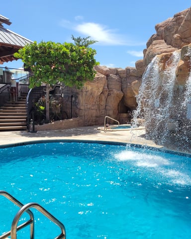 Resort pool with waterfall feature and rock grotto, Tiki Bar pavilion visible in the background under blue sky