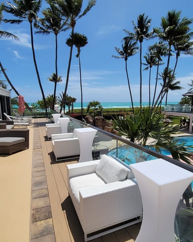 Cabana upper balcony deck with white lounge chairs, pool below, palm trees, and turquoise ocean in the background