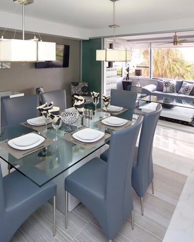 Open-plan dining and living room with glass table, gray upholstered chairs, purple sofa, and large windows overlooking palm trees.