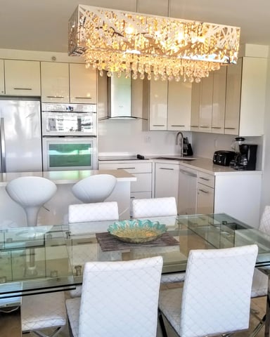 Modern open-plan kitchen and dining area with white cabinetry, stainless steel appliances, glass dining table, and crystal chandelier overhead.