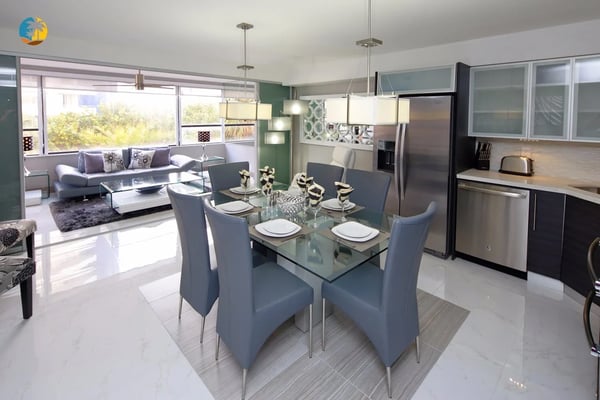 Open-plan kitchen and dining area with glass table, blue chairs, stainless steel appliances, and living room with sofa visible beyond.