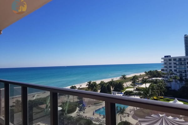 Oceanfront balcony view with turquoise water, sandy beach, palm trees, and beachfront resort buildings