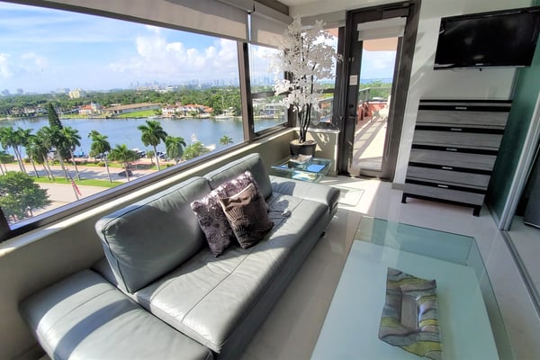 Modern living room with gray sofa, floor-to-ceiling windows overlooking river, palm trees, and distant city skyline