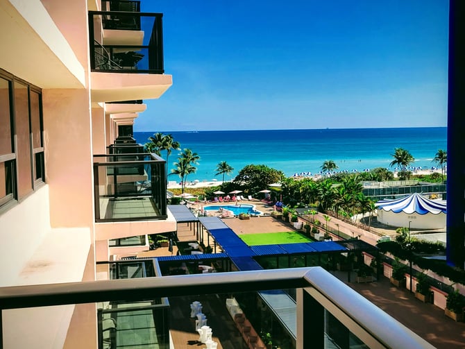 Vista desde el balcón de un complejo turístico frente a la playa, con mar turquesa, playa de arena, palmeras, piscina y sombrillas a rayas