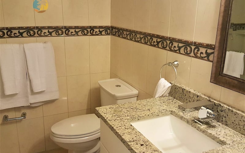 Bathroom with cream tile walls, decorative border trim, granite countertop, white sink, toilet, and towel rack with white towels