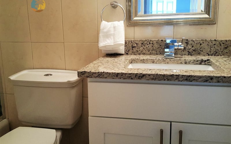 Bathroom with white toilet, speckled granite vanity, sink, mirror, and window view of blue building exterior.