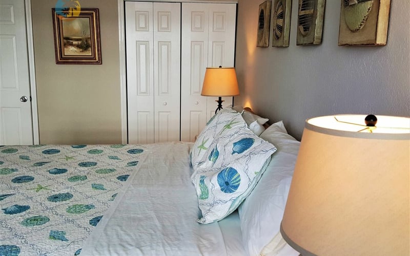 Bedroom with queen bed, patterned pillows, orange table lamp, white closet doors, and framed wall art