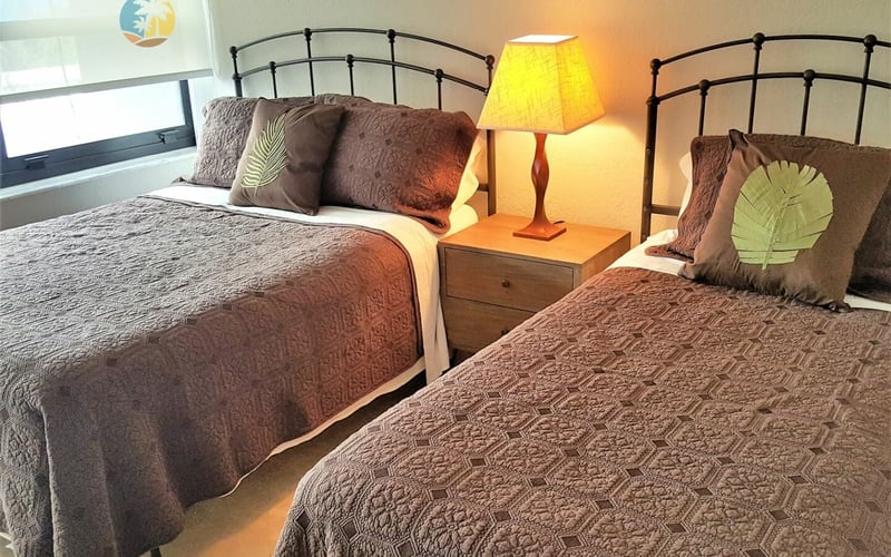 Bedroom with two double beds, black metal headboards, patterned bedspreads, decorative pillows, and a shared nightstand with yellow lamp between them.