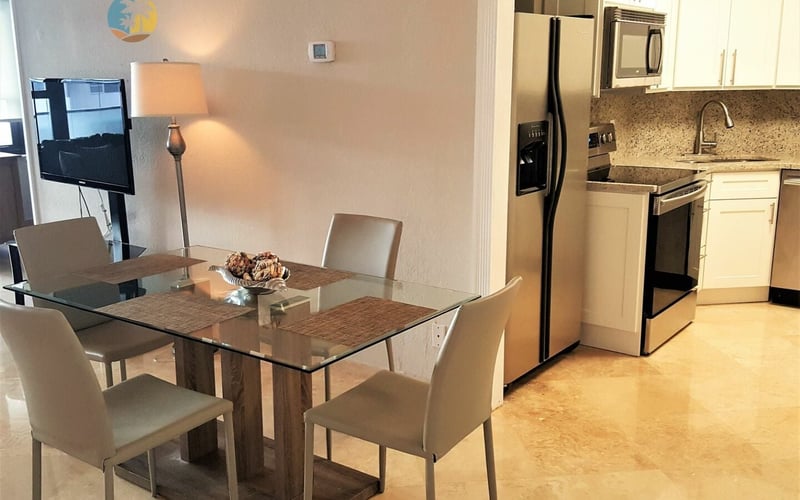 Open-plan kitchen and dining area with glass-top table, beige chairs, stainless steel appliances, and granite countertops.