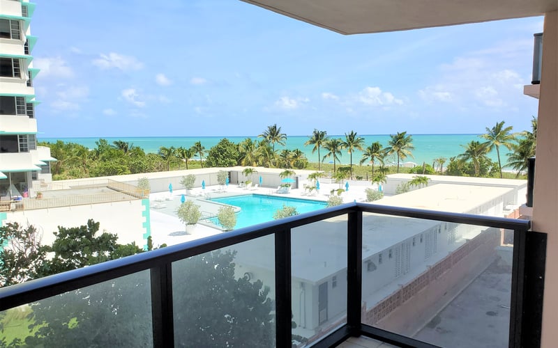 Balcony view of turquoise ocean, palm trees, and resort pool with white deck and blue umbrellas