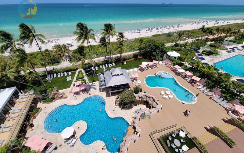Aerial view of resort with multiple pools, palm trees, sandy beach, and turquoise ocean with swimmers