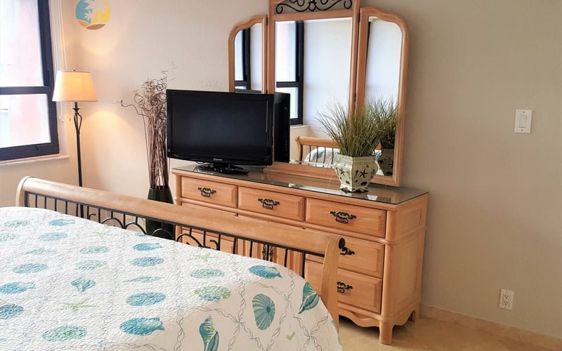 Bedroom with queen bed, wooden dresser with TV, ornate mirror, and lamp beside windows