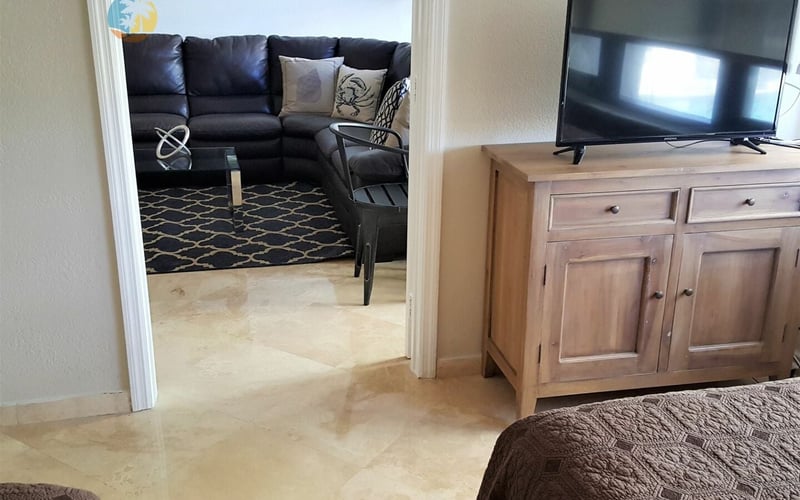 Bedroom with open doorway to living room featuring black leather sectional sofa and flat-screen TV on wooden dresser