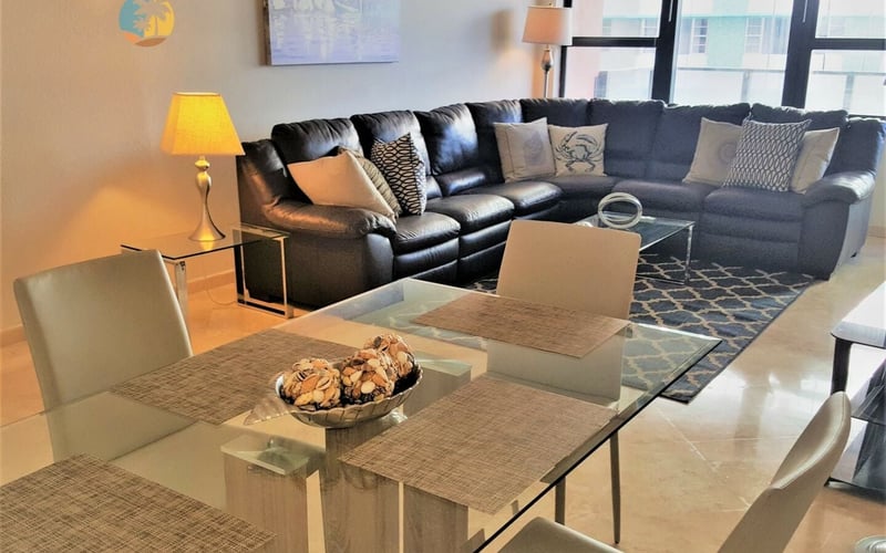 Modern living room with black sectional sofa, glass dining table, table lamp, and city views from floor-to-ceiling windows