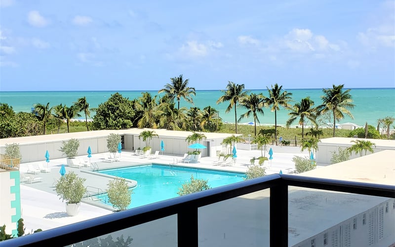 Resort pool with turquoise water, blue umbrellas, and palm trees overlooking tropical ocean and beach
