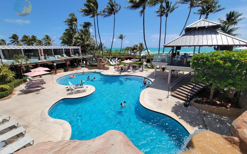 Resort pool with turquoise water, palm trees, beach pavilion, and ocean view beyond