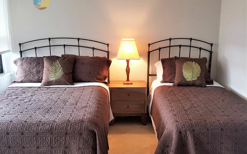 Bedroom with two twin beds with metal headboards, brown quilts, decorative pillows, and a shared nightstand with lamp between them.