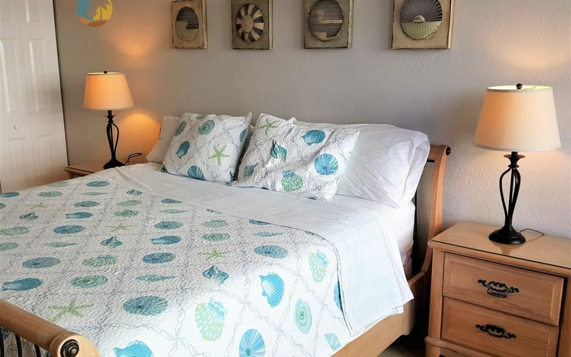 Bedroom with wooden bed, seashell-patterned bedding, two nightstands with orange lamps, and four framed shell artworks on wall